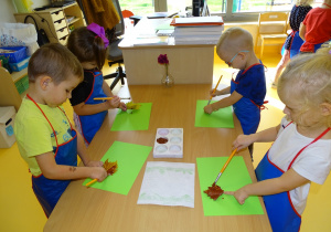 Dzieci stoją przy stoliku, malują liście brązową farbą
